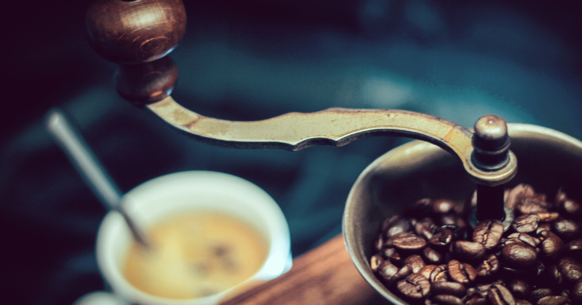 Manual coffee grinder in foreground with mug of fresh coffee