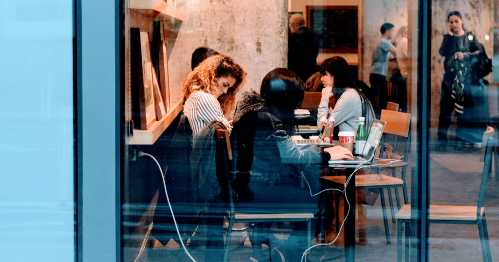 Looking in through a coffee shop window one sees people at a table with open laptops and cups of coffee