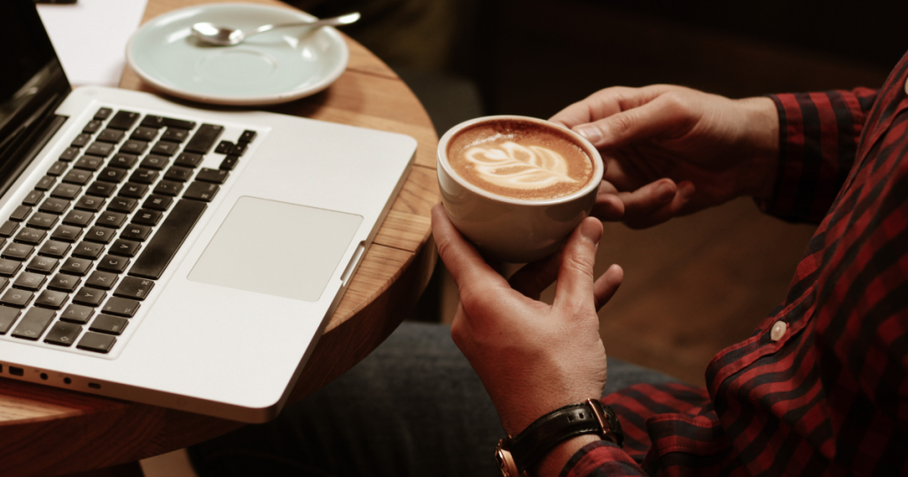A person holding a cup of coffee in their hands at a coffee shop table with an open laptop on the table.